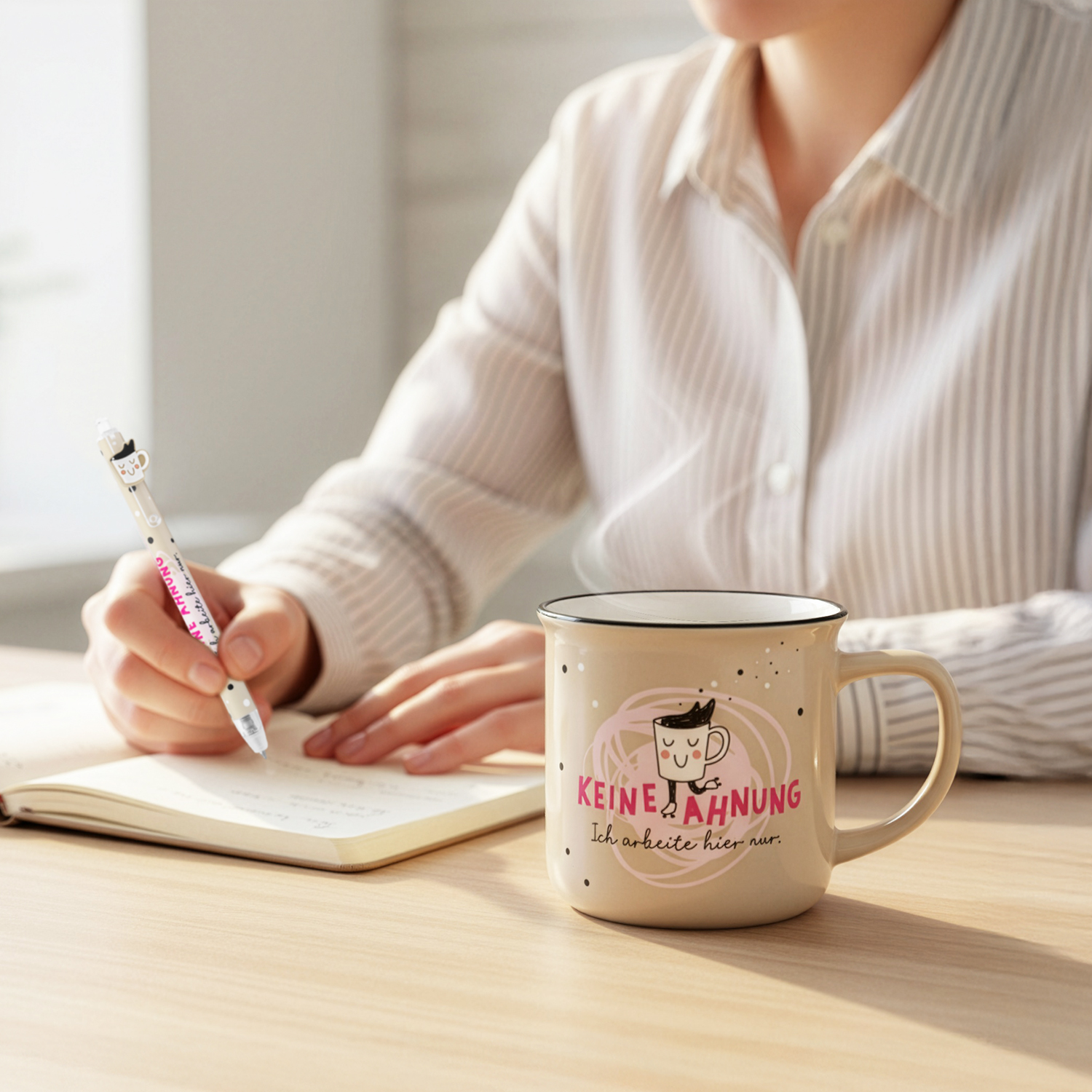 Beige Keramiktasse mit rosa Druck 'KEINE AHNUNG' und Cartoon-Motiv; steht neben Notizbuch.