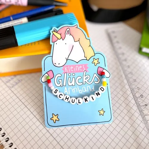 Ein kleines Armband mit der Aufschrift "Schulkind", angebracht an einer Headerkarte mit Einhorn-Motiv.