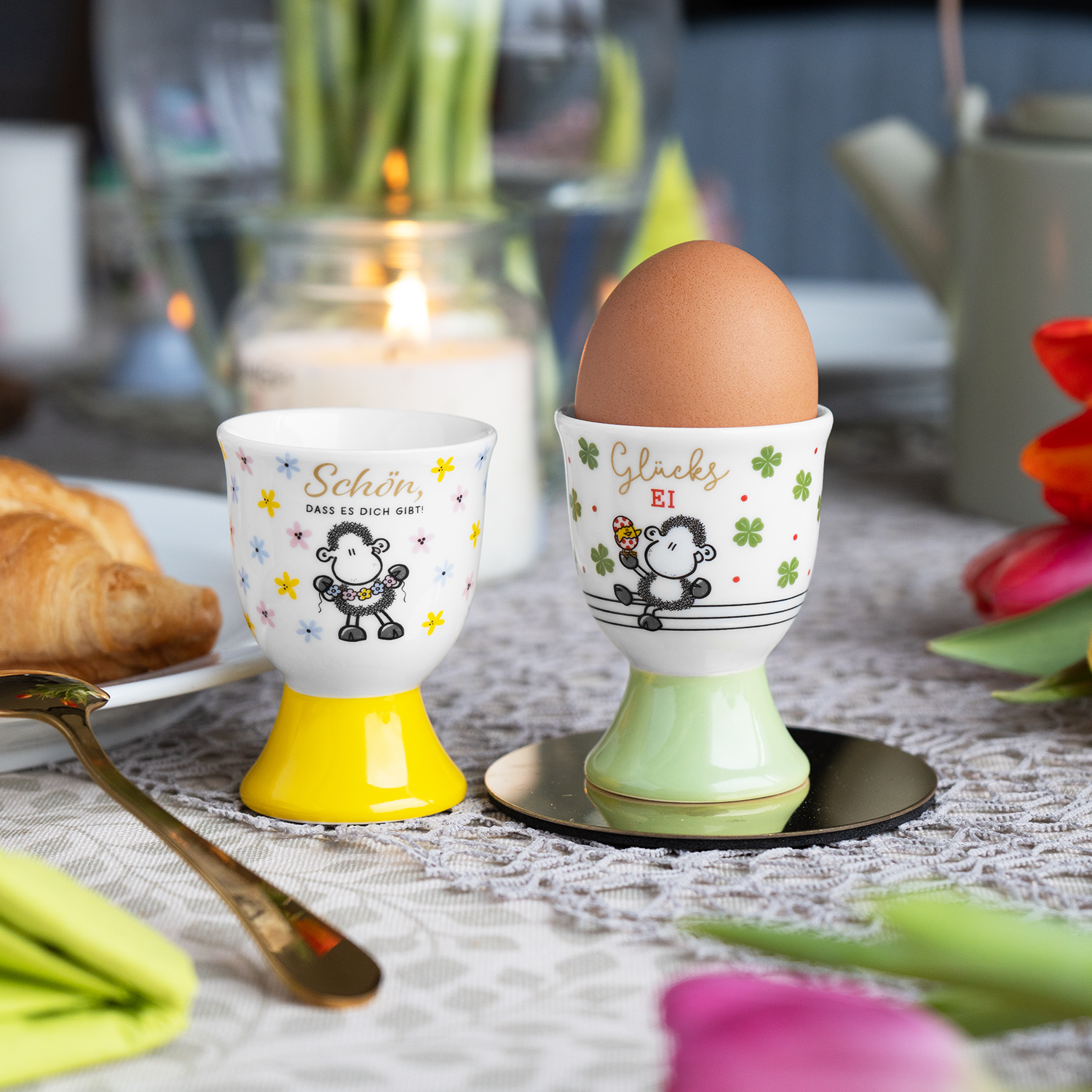 Zwei Porzellan-Eierbecher mit Schaf-Motiven, bunten Blumen und Gelb/Mintbasen.