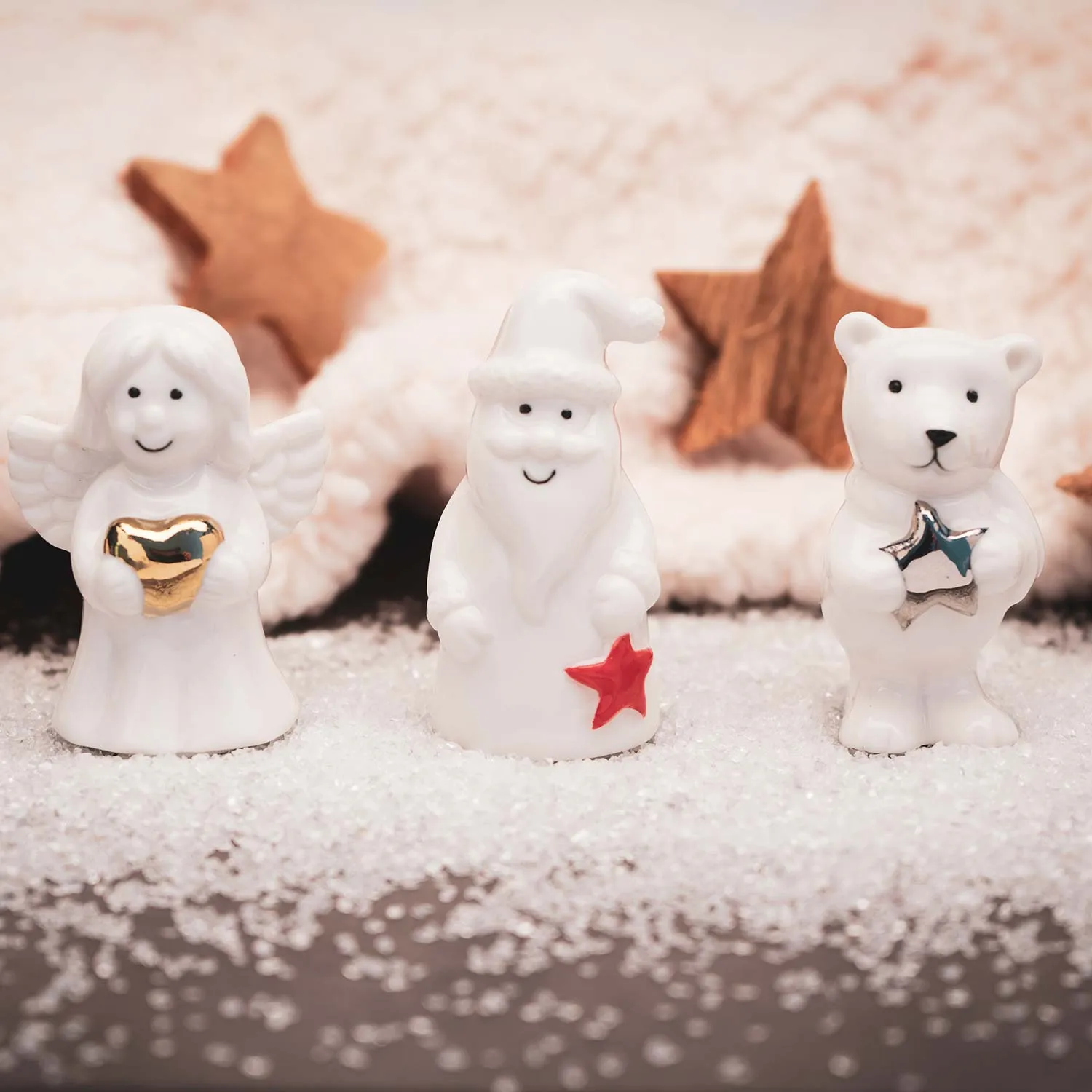 Drei Porzellan-Weihnachtsfiguren – Engel, Nikolaus und Bär – mit goldenem Herz und Sternen auf schneebedeckter Fläche.