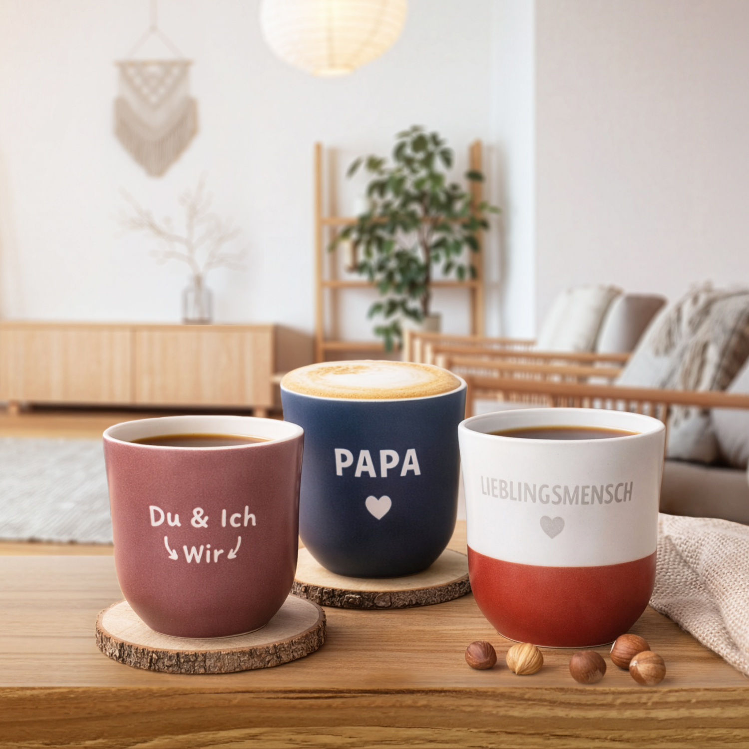 Drei Keramikbecher mit Sprüchen in Pink, Blau und Weiß-Rot, stehen auf Holzuntersetzern