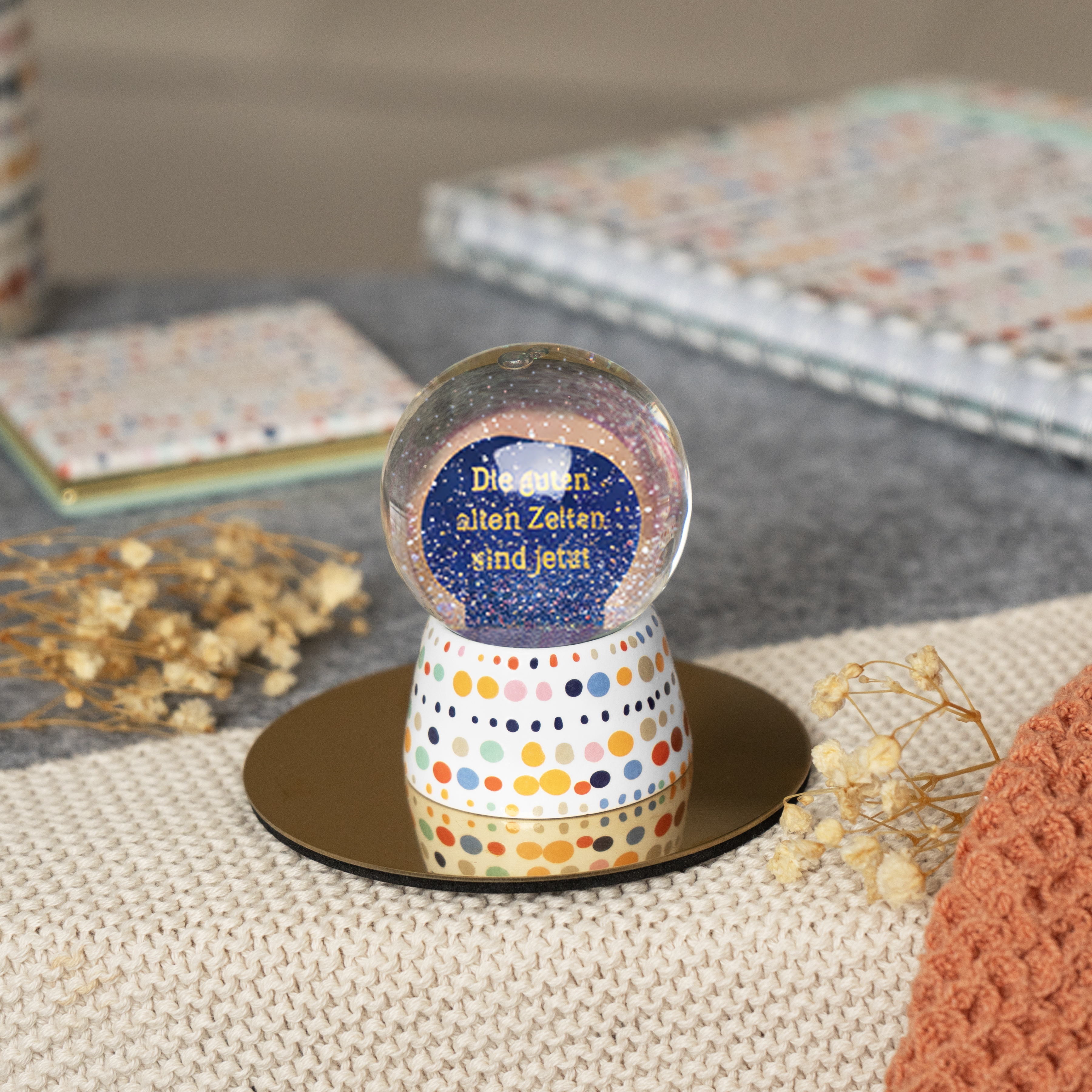 Schneekugel »Zeiten« mit blauem Glitter, buntem Sockel und gelbem Text.