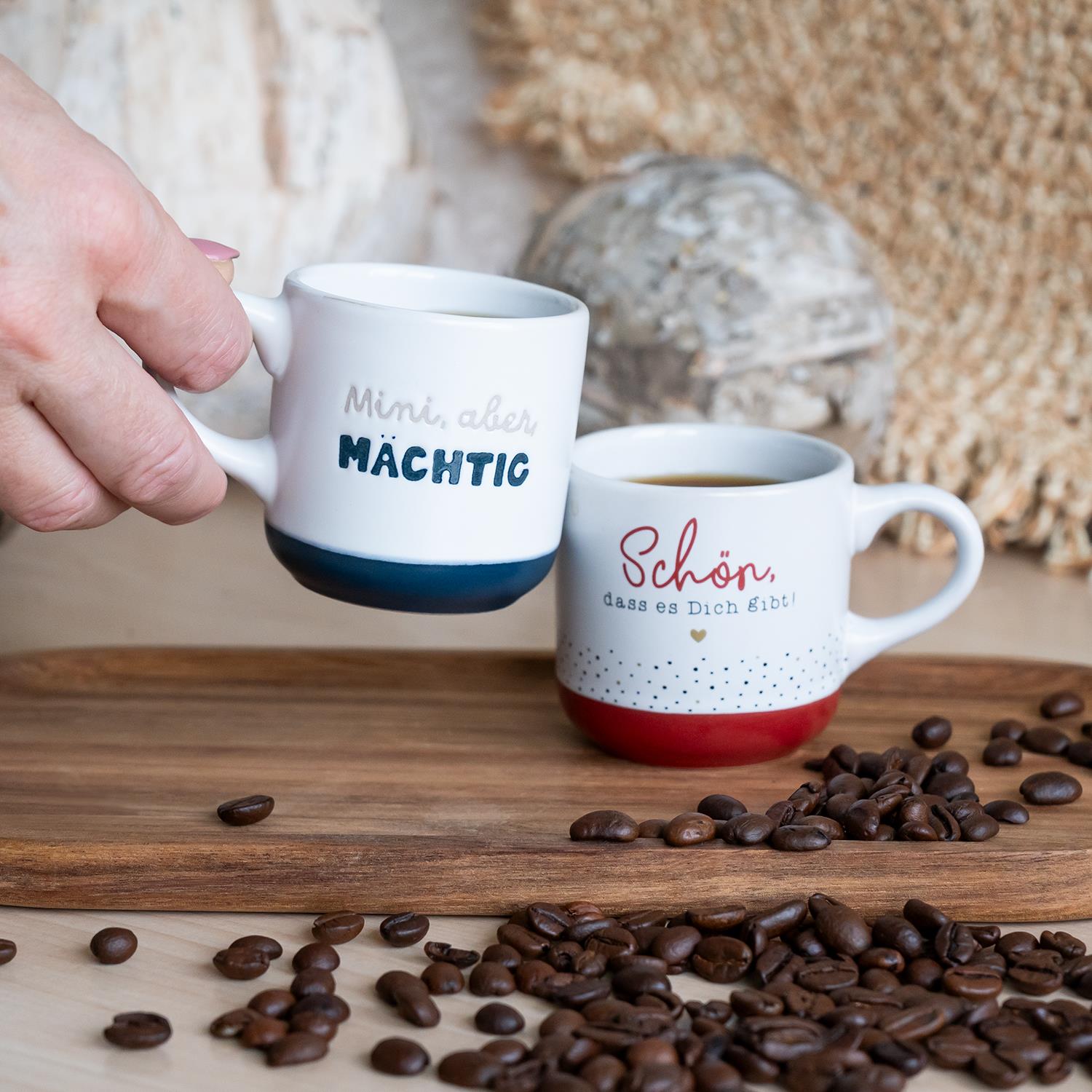 Zwei weiße Espressotassen aus Steinzeug mit Sprüchen auf farblich abgesetztem Boden (blau und rot) auf Holzunterlage.