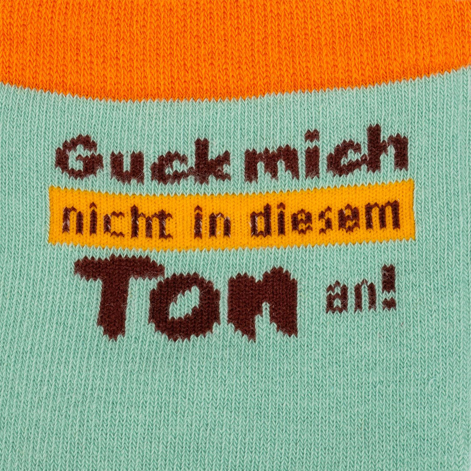 Mintgrüne Socke mit orangefarbenem Streifen und gelbem Label mit dem Spruch 'Guck mich an' in braunen Buchstaben.