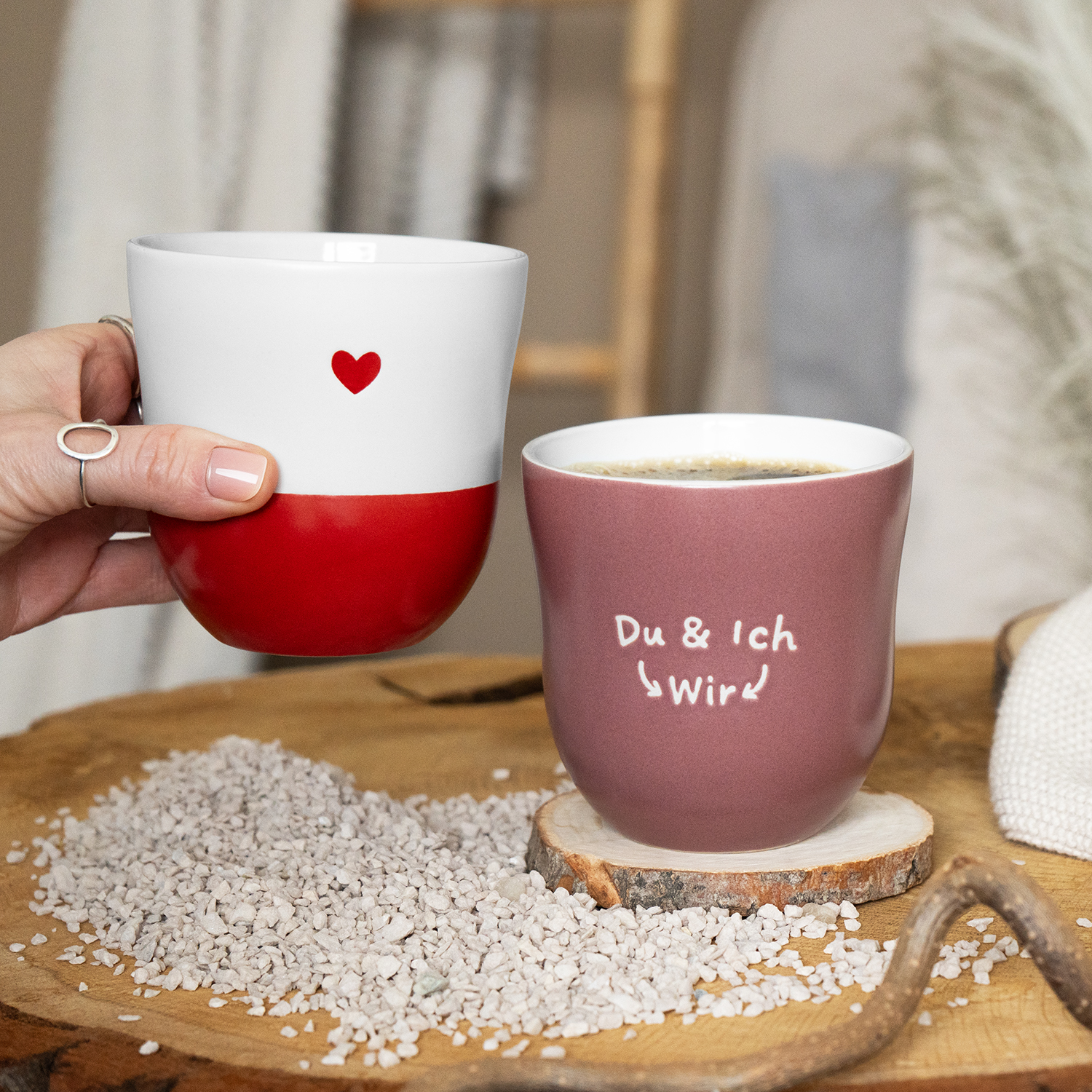 Zwei Keramik-Becher: Weißer Becher mit rotem Unterteil und Herz, rosafarbener Becher mit Schriftzug Du & Ich → Wir